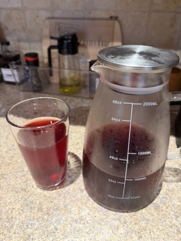 A glass of deep pink hibiscus lemonade next to a full glass pitcher on a kitchen counter