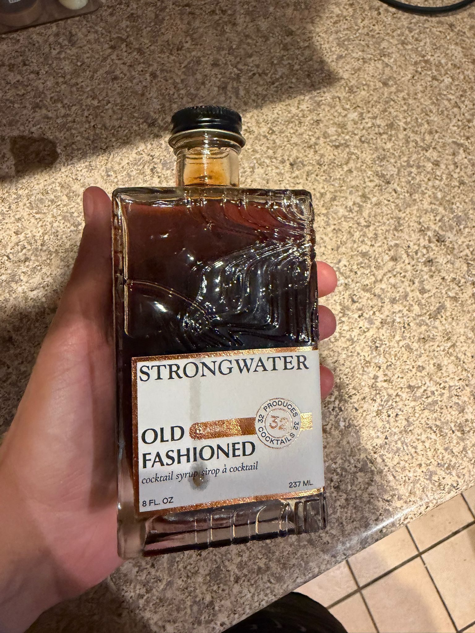 My hand holding a bottle of Strongwater Old Fashioned cocktail syrup on the kitchen counter